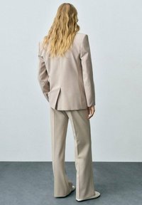 Blazer beige sur mesure avec une seule fente, associé à un pantalon large assorti. Tissu lisse avec un design formel.