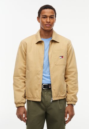 Joven con una chaqueta beige con cremallera, camisa azul clara, pantalones verdes y un cinturón negro con hebilla rectangular.