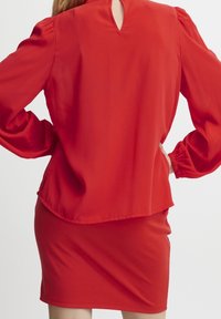 Rote Bluse mit langen Ärmeln und einem Schlüsselaugen-Detail am Rücken, kombiniert mit einem enganliegenden roten Rock. Beide Kleidungsstücke haben einen glatten Stoff und eine elegante Silhouette.