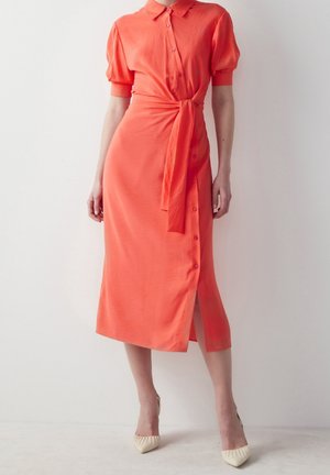 Woman wearing a coral midi dress with puff sleeves, buttoned front, waist tie, and side slit paired with cream pointed heels.