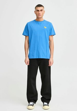 Jaunas vyras stovi, vilkėdamas ryškiai mėlyną marškinėlį su mažu geltonu logotipu, juodas plačias kelnes ir juodai-baltus sportbačius.