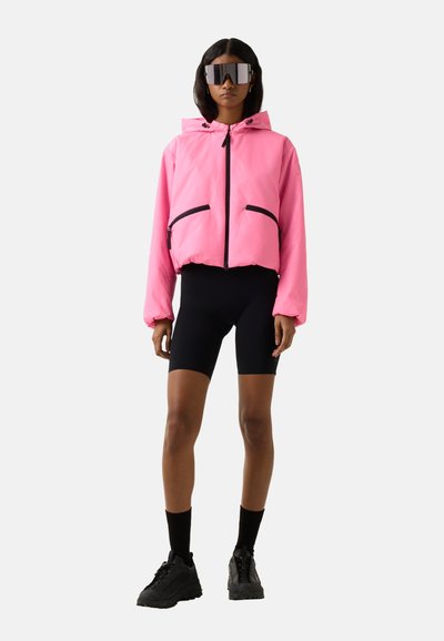 Frau trägt übergroße pinke Kapuzenjacke, schwarze Radlerhosen, schwarze Socken, klobige schwarze Sneakers und große reflektierende Sonnenbrille, steht vor weißem Hintergrund.