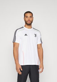 Biała sportowa koszulka z krótkimi rękawami, czarnymi paskami na ramionach, zielonymi akcentami i czerwonymi szwami po bokach, z logo Juventusu i brandingiem Adidas.