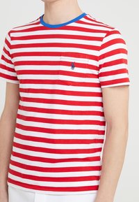 Červené a bílé pruhované tričko s modrým límečkem, obsahující náprsní kapsu a vyšívané logo. Krátké rukávy, bavlněný materiál, uvolněný střih.