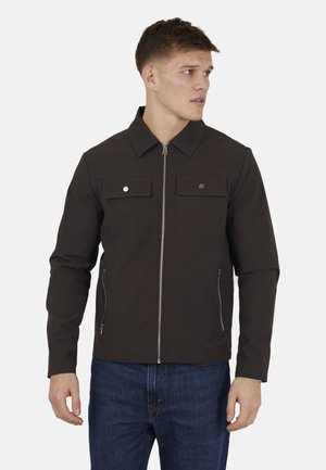 Młody mężczyzna ubrany w ciemnobrązową kurtkę zapinaną na zamek z klapowymi kieszeniami na wysokości klatki piersiowej i kieszeniami bocznymi na zamek, w połączeniu z niebieskimi dżinsami, patrzący w prawo.