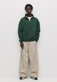 Grøn fleece-pullover med en kvart lynlås, parret med oversized lys beige denimjeans. Fodtøjet har sorte og sølvfarvede accenter.