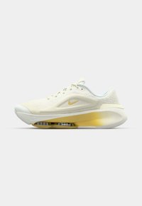 Nike sneaker met een gestructureerde lichtromige bovenkant, gele luchtkussens en een witte rubberen zool. Heeft een klein goudkleurig logo aan de zijkant.