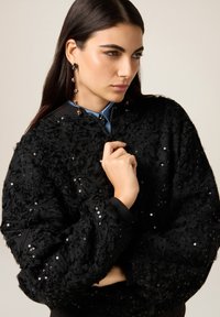 Giacca bomber nera con trama a rilievo e superficie di paillettes, chiusura frontale con zip e polsini a coste. Camicia blu con colletto sotto, caratterizzata da un colletto con bottoni.