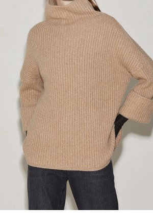 Beige geribbelde gebreide trui met een hoge kraag, voorzien van losse mouwen en zijsplitten, gecombineerd met donker gestreepte jeans.