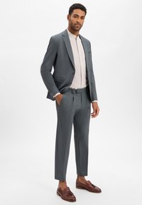 Grijs op maat gemaakt kostuum met een gestructureerde afwerking, bijpassende broek en een lichtbeige overhemd. Bruine loafers maken de look af.