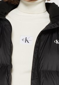 Černá prošívaná vesta na zip s měkkou černou podšívkou, nošená přes bílý žebrovaný rolák s logem Calvin Klein Jeans.