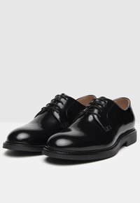 Scarpe da uomo in pelle nera con finitura lucida, punta tonda, chiusura con lacci e dettagli cuciti lungo la suola. Suola in gomma piatta.