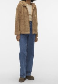 Tann fuzzy jacka med dragkedja framtill och huva, bärs över en ljusbeige topp, ihop med blå vida jeans och bruna klumpiga stövlar.