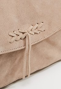 Beige semišová taška s přehnutým klopou, zdobená dekorativním šitím a zavázaným semišovým akcentem. Hladký povrch a minimalistický design.