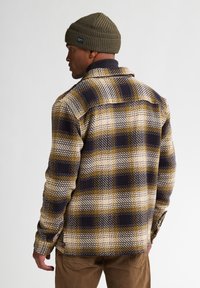 Geruite flanellen blouse in geel, marineblauw en crèmekleur; met knopen en een kraag, van textuurstof. Draagt een groene gebreide beanie.