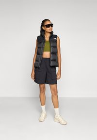 Svart pufferväst med hög krage, olivgrönt crop top och svarta cargoshorts. Vita sneakers och solglasögon fulländar outfiten.