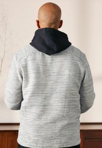 Grå tekstureret sweater med en marineblå hætte, der har vandrette linjer, lange ærmer og en afslappet pasform. Set bagfra, ingen yderligere detaljer.