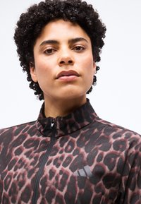 Personne aux cheveux courts et bouclés portant une veste à col montant imprimée léopard avec un logo Adidas visible, regardant la caméra avec confiance.