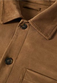 Brun ruskindskjorte med spidse krave, to sorte knapper og en brystlomme. Blød tekstur og glat overflade med syede detaljer.