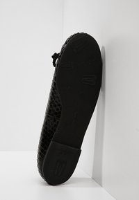 Semelle d’une chaussure noire texturée avec des motifs en relief et un détail de lacet, placée verticalement contre un fond blanc en coin.
