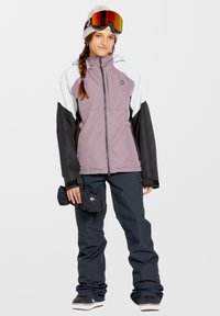 Snowboardjack in lavendel, zwart en wit kleurblokdesign. Voorzien van een ritssluiting en een waterdicht materiaal. Gecombineerd met donkere sneeuwbroeken.