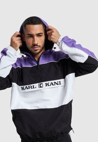 Črna, bela in vijolična vetrovka s kapuco in zadrgo, ki ima spredaj logo in kapuco s vrvico. Lahka, športna oblika.