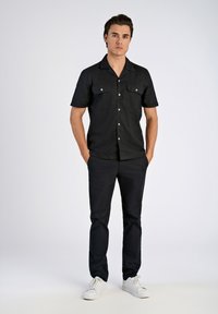 Chemise noire à manches courtes avec deux poches poitrine et boutons argentés, associée à un pantalon noir et des baskets blanches sur un fond neutre.