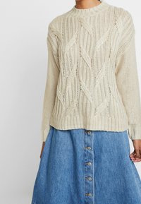 Pull beige en maille torsadée à manches longues, associé à une jupe en denim bleue boutonnée à l'avant.