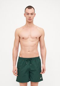 Grønne svømmeshorts laget av lett materiale, med elastisk midjebånd og snøring, samt en liten merkeplakat på siden.