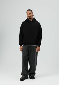 Schwarzer Kapuzenpullover mit einer Fronttasche, kombiniert mit dunkelgrauen Jeans mit weitem Bein. Die Schuhe haben eine klobige schwarze Sohle und bunte Schnürsenkel.