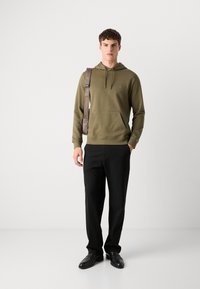 Olijfgroene hoodie met een kangoeroezak, gecombineerd met zwarte broek en zwarte schoenen, met een eenvoudig ontwerp en subtiele branding.