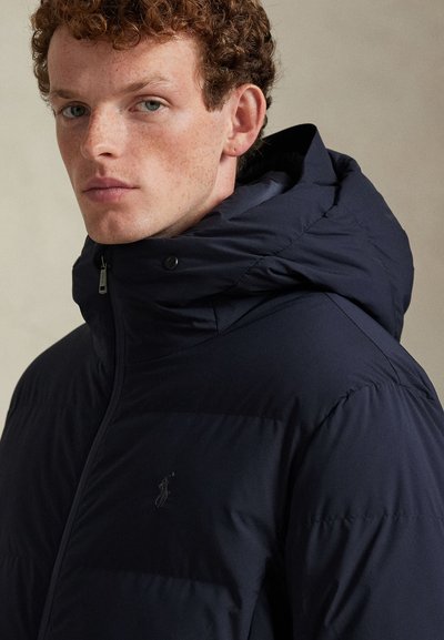 Tamsiai mėlyna pūsta striukė su aukšta apykakle, užtrauktuku ir spaudėmis. Turi horizontalius siuvimus ir toninį logotipą ant krūtinės.