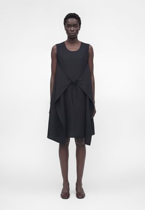 Kvinde står forfra iført en ærmeløs sort kjole med asymmetrisk overlag bundet i taljen og mørke flade sko på hvid baggrund.