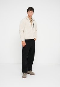 Uomo che indossa una giacca in pile beige, una camicia beige, pantaloni neri a gamba larga e stivali grigi, in piedi di fronte a uno sfondo bianco uniforme.
