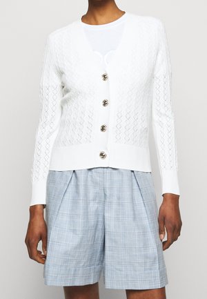 Cardigan blanc texturé boutonné associé à un short plissé à carreaux bleu clair porté par une personne à la peau brune.