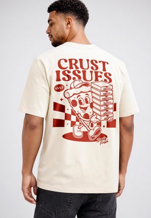 Mann trägt cremefarbenes T-Shirt mit roter Grafik einer Cartoon-Pizzascheibe, die gestapelte Pizzakartons hält, beschriftet mit Wochentagen und Text "Crust Issues."