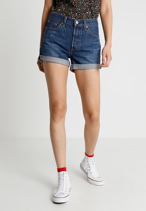 Mujer con shorts vaqueros azules remangados, zapatillas blancas de caña alta, calcetines rojos hasta el tobillo y una blusa oscura con estampado floral, posando frente a un fondo liso.