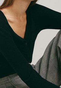 Haut noir côtelé avec un décolleté en V et un détail d'un seul bouton, associé à un pantalon taille haute à carreaux gris. Texture lisse, coupe ajustée.