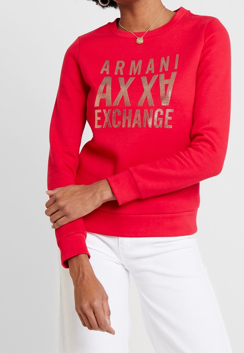 Femme portant un sweat-shirt rouge Armani Exchange avec logo doré, pantalon blanc et un collier avec un pendentif rond en or, bras croisés à la taille.