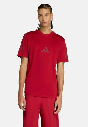 Junger Mann mit geflochtenem Haar, der ein rotes Adidas-T-Shirt und passende rote Shorts trägt, vor einem schlichten weißen Hintergrund stehend.