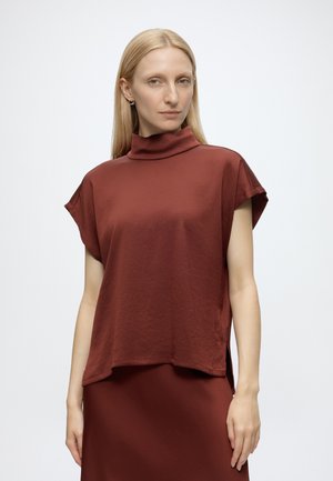 Femme aux cheveux blonds lisses portant un haut sans manches couleur rouille à col montant et une jupe assortie sur un fond uni clair.