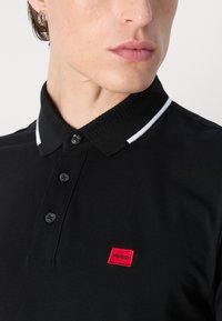 Fekete póló ing, textúrált felszínnel, fehér színű gallér csíkokkal, háromgombos gombolással és egy kis piros hímzett "HUGO" logóval a mellkasán.