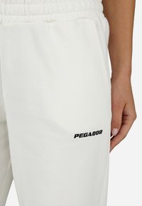 Baltas kokvilnas sporta bikses ar elastīgu jostu un vārdu "PECADOR" melnā izšuvumā uz kreisā augšstilba. Gluda tekstūra.