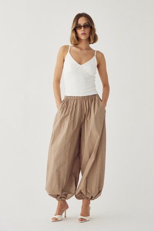 Donna che indossa una canotta bianca senza maniche, pantaloni beige a gamba larga con polsini elastici, sandali bianchi con tacco alto e occhiali da sole scuri, in piedi davanti a uno sfondo bianco.