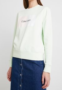 Mátově zelená mikina s kulatým výstřihem a žebrovanými manžetami, s bílým obdélníkovým motivem s nápisem "Calvin Klein". Kombinováno s džínovou sukní.