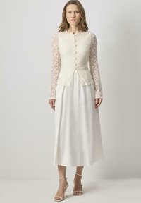 Lace and satin dress in cream with a fitted bodice and long lace sleeves. The skirt flows to ankle length, featuring a modest neckline and buttons.