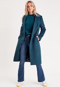 Trinchera de mezcla de lana en color verde azulado con cuello con muesca, dos botones frontales y bolsillos laterales, superpuesta sobre un suéter texturizado y unos vaqueros acampanados.