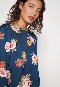Blusa floral com fundo azul, decorada com rosas cor-de-rosa e laranja. Tecido suave, decote redondo e mangas ligeiramente bufantes.