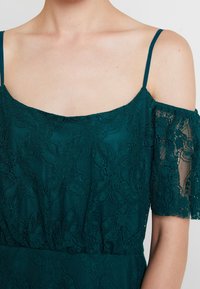 Tyrkysové krajkové šaty s odkrytými rameny, květinovým vzorem a jemnou texturou. Tenké nastavitelné ramínka dotvářejí design.