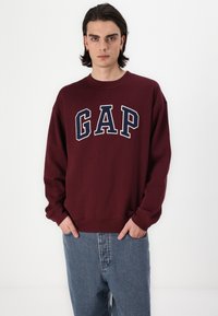 Marinröd sweatshirt med "GAP" tryckt i vita och marinblå bokstäver. Har rundad halsringning och elastiska mudd. Matchas med ljusblå jeans.